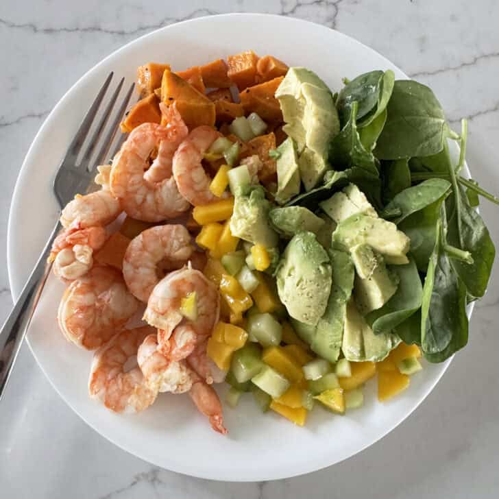 White plate with shrimp, mango, cucumbers, spinach, avocado and sweet potatoes.