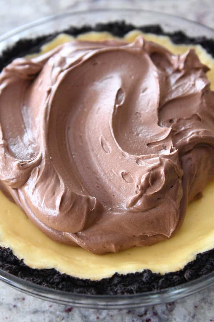 Whipped chocolate mousse getting spread over a cheesecake with an Oreo crust in a glass pie dish.