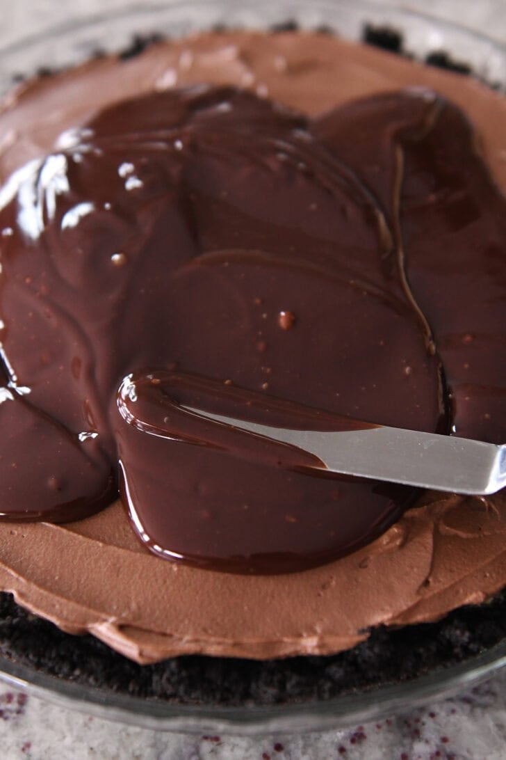 Chocolate ganache being spread with a metal spatula over a chocolate mousse cheesecake in a glass pie dish.