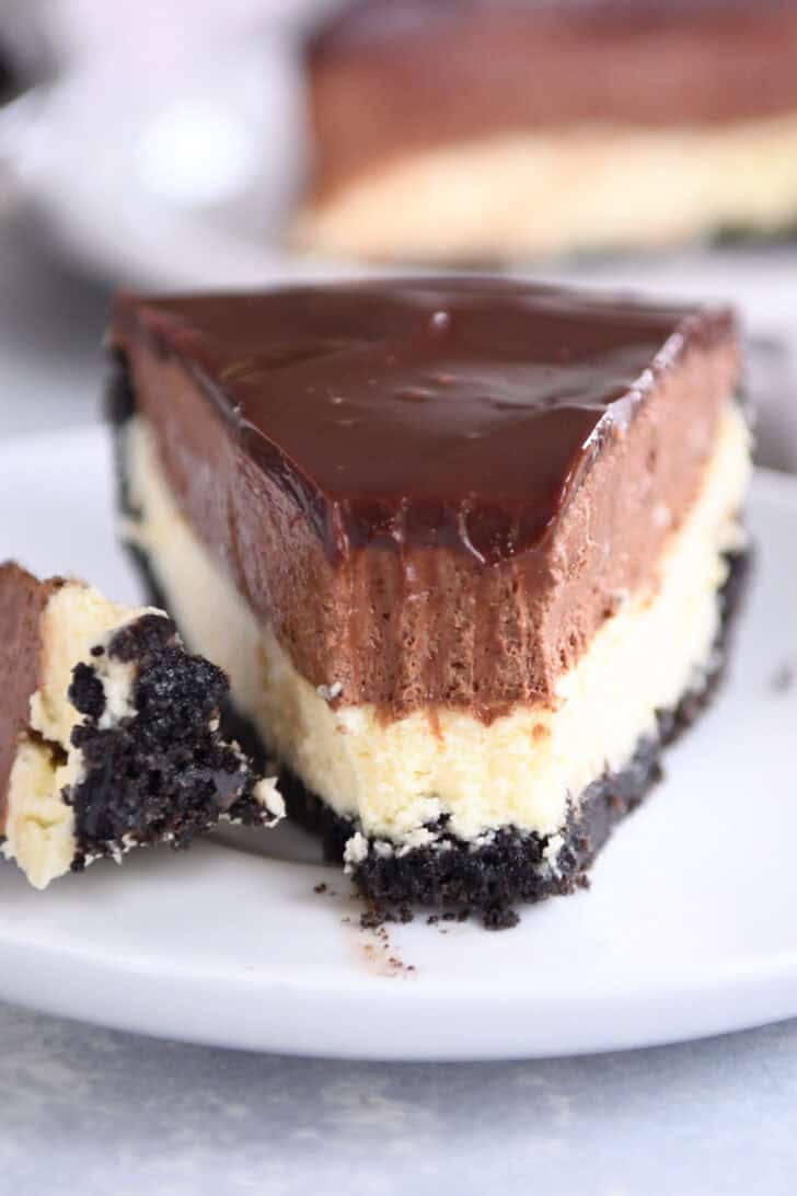 A white plate holding a slice of Oreo crust cheesecake, layered with creamy cheesecake, rich chocolate mousse, and topped with smooth chocolate ganache. A bite is taken out of the slice.