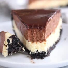 A white plate holding a slice of Oreo crust cheesecake, layered with creamy cheesecake, rich chocolate mousse, and topped with smooth chocolate ganache. A bite is taken out of the slice.