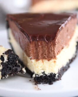 A white plate holding a slice of Oreo crust cheesecake, layered with creamy cheesecake, rich chocolate mousse, and topped with smooth chocolate ganache. A bite is taken out of the slice.