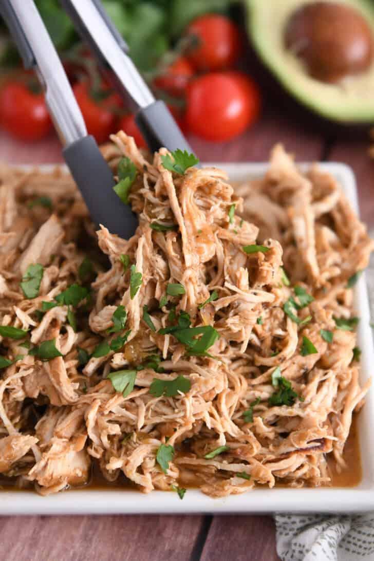 Tongs grabbing portion of salsa verde chicken on white tray.