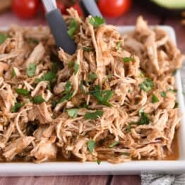 Tongs grabbing portion of salsa verde chicken on white tray.