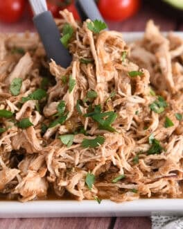 Tongs grabbing portion of salsa verde chicken on white tray.