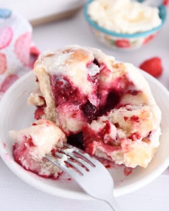 Fork taking piece of raspberry sweet roll on white plate.