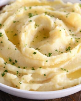 A white bowl with buttery mashed potatoes topped with parsley flakes. A wooden spoon is in the bowl.