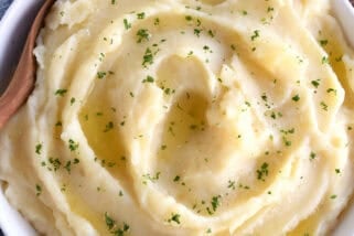 Top down view of a white bowl with buttery mashed potatoes topped with parsley flakes. A wooden spoon is in the bowl.