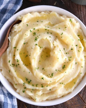 Top down view of a white bowl with buttery mashed potatoes topped with parsley flakes. A wooden spoon is in the bowl.