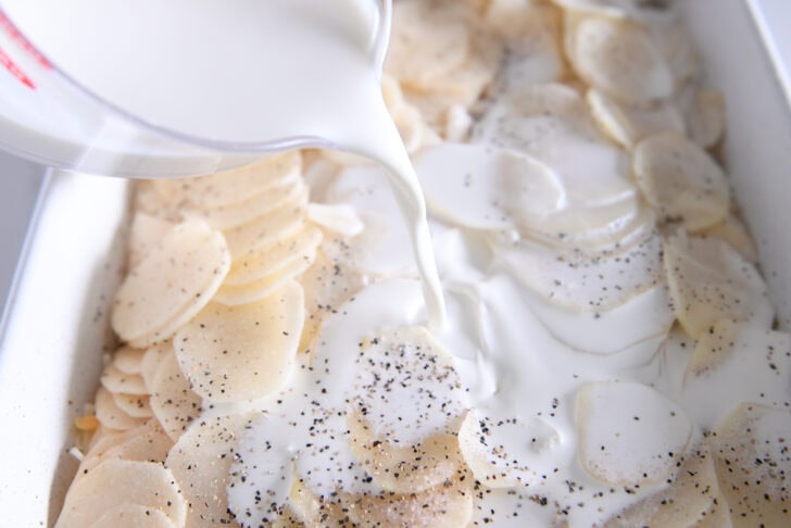 Pouring cream over thin slices of potatoes in white 9X13-inch dish.