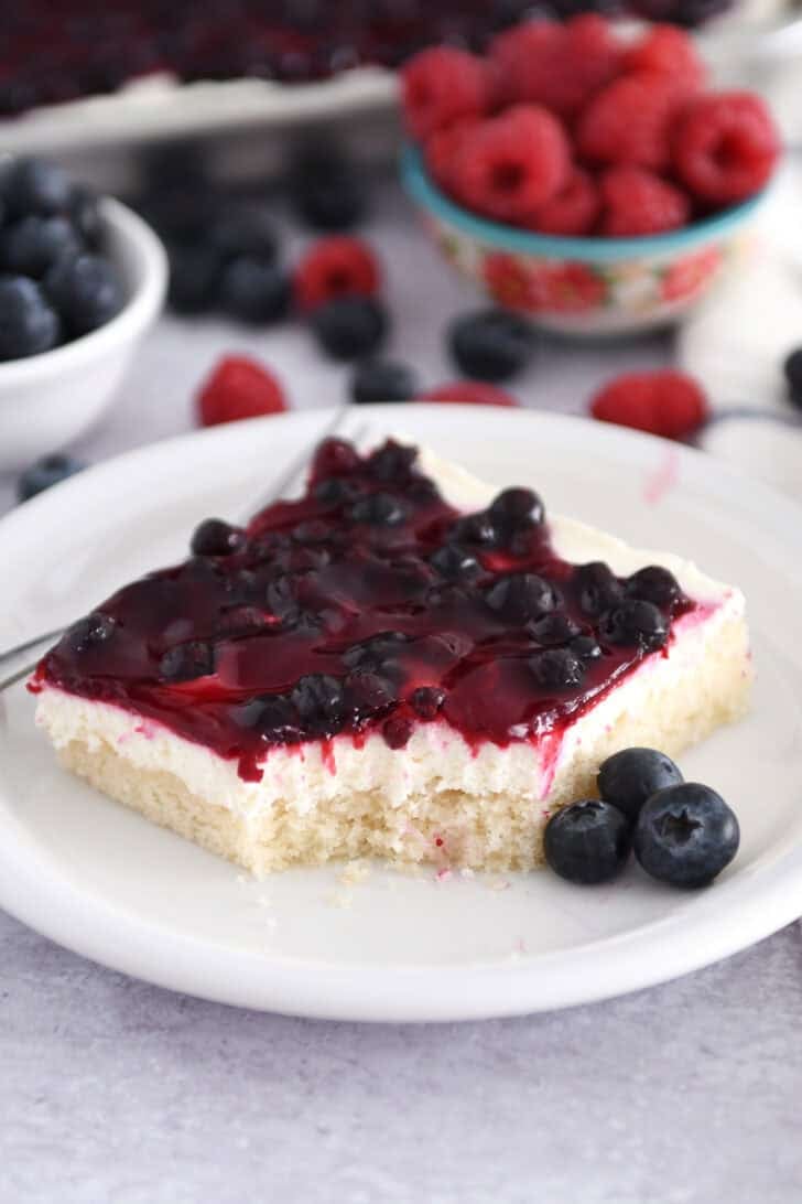 Square of white sheet cake topped with cream cheese layer and fruit topping on white plate with fresh blueberries.