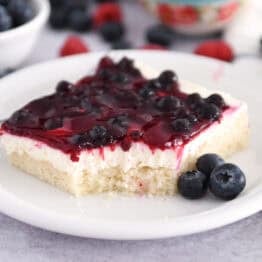 Square of white sheet cake topped with cream cheese layer and fruit topping on white plate with fresh blueberries.