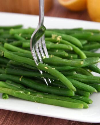 Fork piercing several fresh, cooked green beans on white tray.