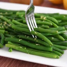 Fork piercing several fresh, cooked green beans on white tray.