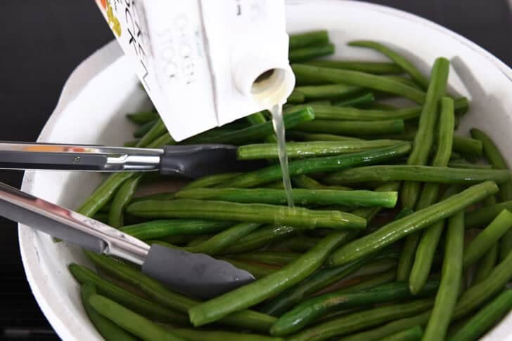 Pouring chicken broth into skillet with fresh green beans and tongs.