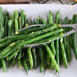 White tray with cooked green beans, lemon zest and tons scooping up portion of green beans.