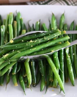 White tray with cooked green beans, lemon zest and tons scooping up portion of green beans.
