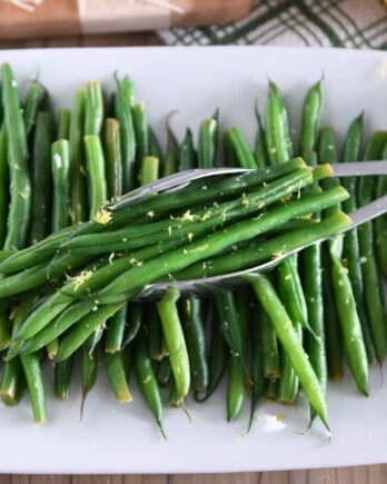 White tray with cooked green beans, lemon zest and tons scooping up portion of green beans.