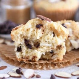 One almond coconut chocolate chip muffin broken in half on brown paper liner.