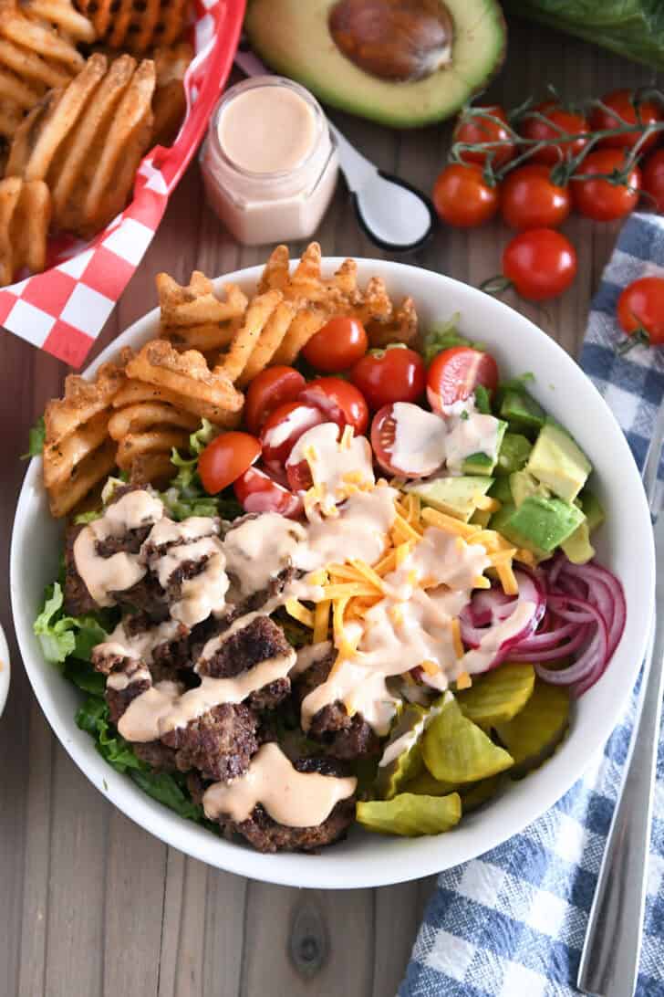 Cooked ground beef, pickles, lettuce, pickled red onions, tomatoes, fries and creamy sauce in white bowl.