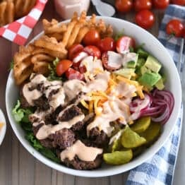 Cooked ground beef, pickles, lettuce, pickled red onions, tomatoes, fries and creamy sauce in white bowl.