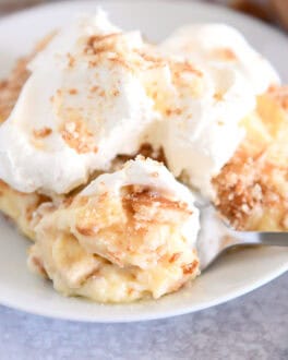 Serving of banana pudding on white plate topped with whipped cream and bite on spoon.