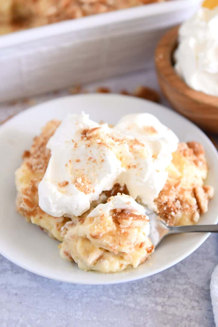 Serving of banana pudding on white plate topped with whipped cream and bite on spoon.