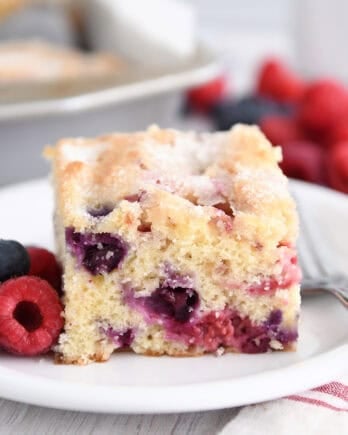 Square of cake with blueberries and raspberries on white plate.