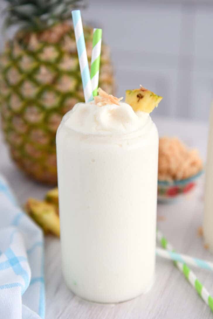 Tall glass cup with frozen piña colada, toasted coconut, a triangle of fresh pineapple and a bit of toasted coconut on top.