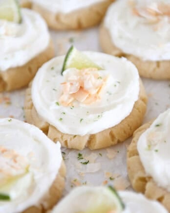 Lots of frosted coconut lime sugar cookies topped with toasted coconut and a fresh lime wedge on white parchment paper.