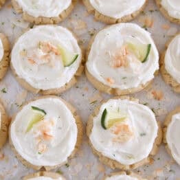 Lots of frosted coconut lime sugar cookies topped with toasted coconut and a fresh lime wedge on white parchment paper.