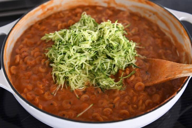 Adding shredded zucchini to cooked pasta in red sauce with shredded zucchini in deep skillet.