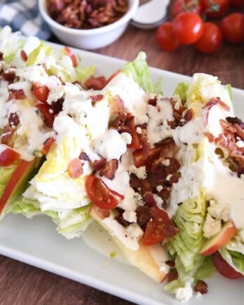 White platter with iceberg wedges, blue cheese crumbles, blue cheese dressing, tomatoes, bacon, and sliced apples.