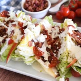 White platter with iceberg wedges, blue cheese crumbles, blue cheese dressing, tomatoes, bacon, and sliced apples.