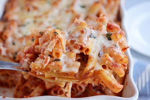 A large metal spoon taking a scoop of cheesy baked ziti out of a casserole dish. 