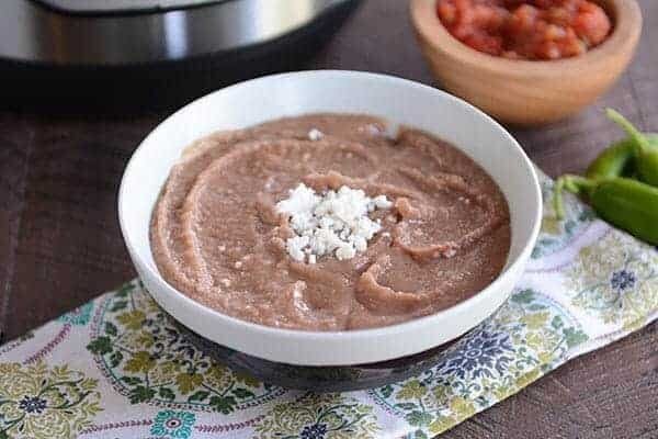 A white bowl of refried beans with a spoon on the side and cheese sprinkled in the middle of the bowl. There is a wooden bowl of chopped tomatoes in the background. 