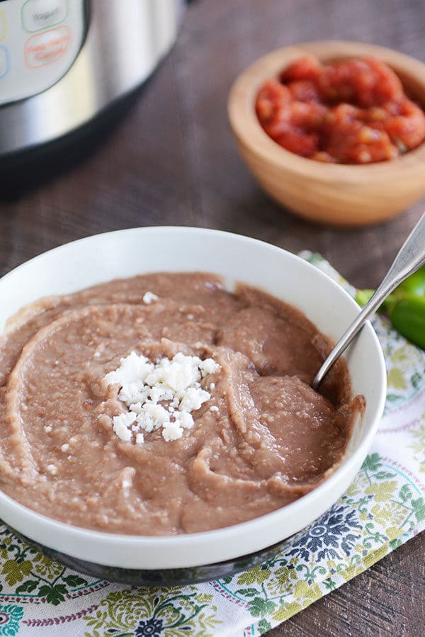 A white bowl of refried beans with a spoon on the side and cheese sprinkled in the middle of the bowl. There is a wooden bowl of chopped tomatoes in the background. 