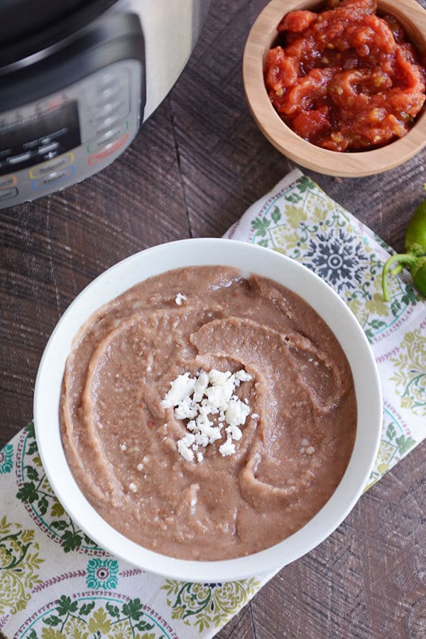 A white bowl of refried beans with a spoon on the side and cheese sprinkled in the middle of the bowl. There is a wooden bowl of chopped tomatoes in the background. 