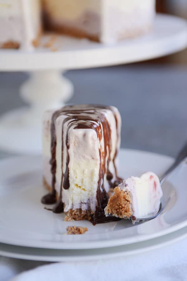 A slice of ice cream cake topped with chocolate syrup on a white plate with one bite of it on a fork next to it.