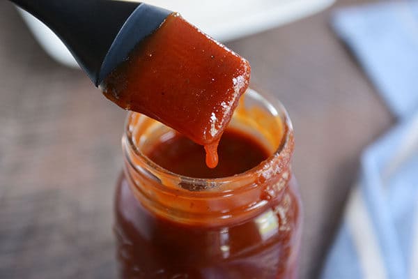 A mason jar full of BBQ sauce, with a drip coming down the side and a pastry brush with sauce over the top.