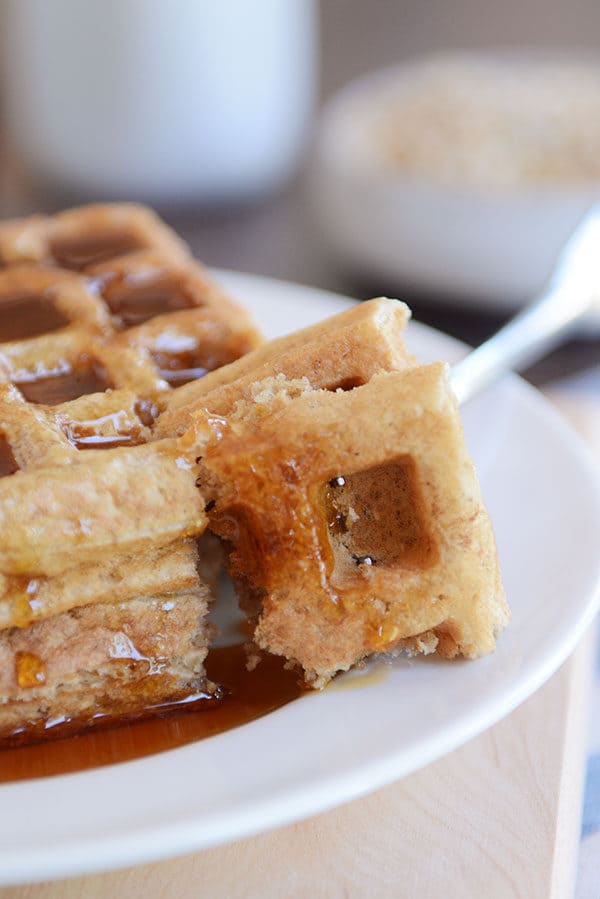 A fork taking a bite out of two thick syrupy waffles on a white plate.