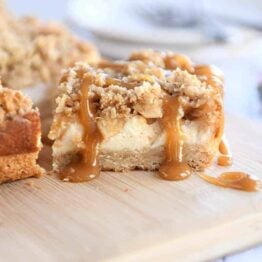 Caramel apple cheesecake bar drizzled with caramel on wood cutting board.