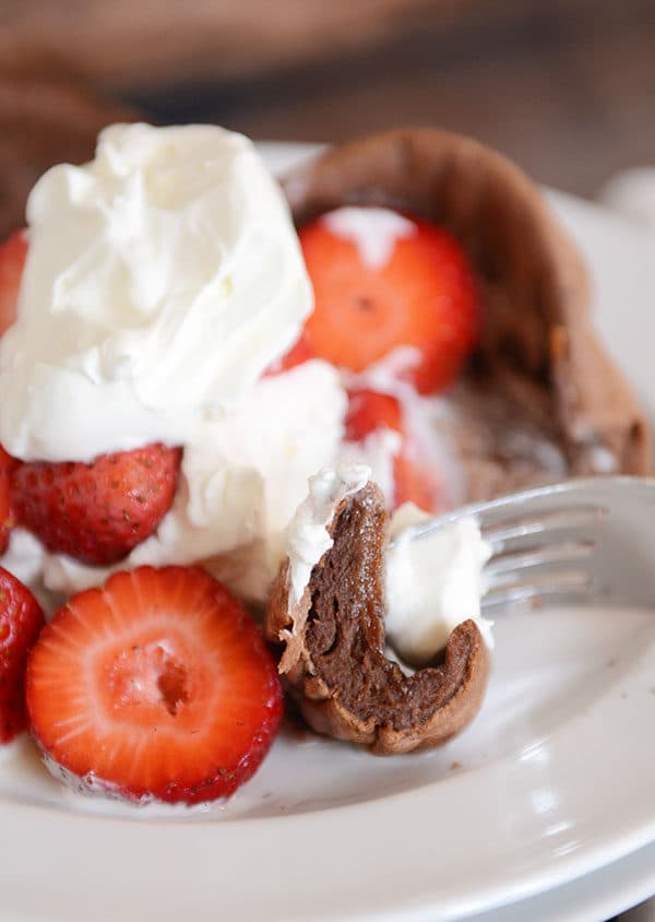 A fork taking a bite out of a chocolate pancake that is covered in whipped topping and sliced strawberries.