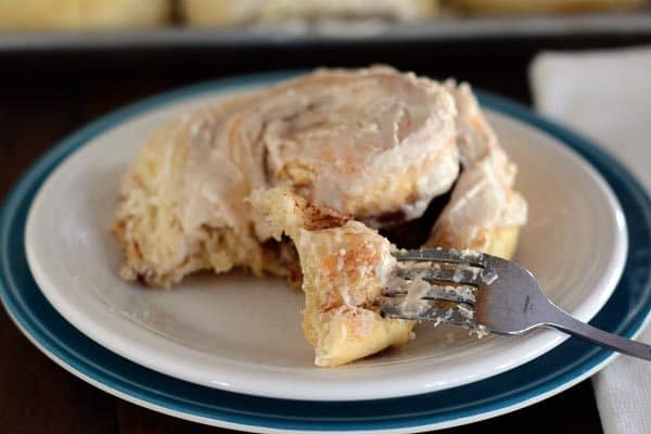 A frosted cinnamon roll with a but being taken out, on a white plate. 