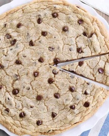 Giant chocolate chip cookie on white platter with slice cut.