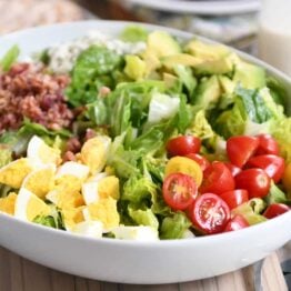 Grilled chicken cobb salad in white bowl with lettuce, tomatoes, eggs, bacon and blue cheese.