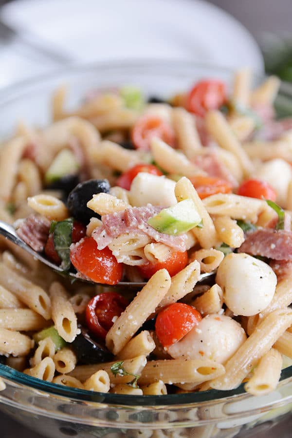A spoon taking a scoop out of Italian pasta salad out of a glass bowl of salad.
