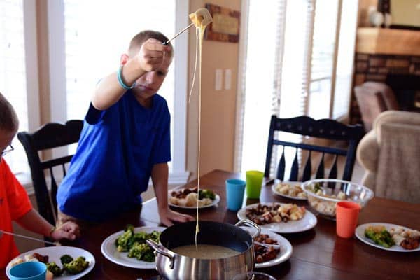 A boy pulling something he dipped out of a fondue pot with a long string of the cheese hanging off the end.