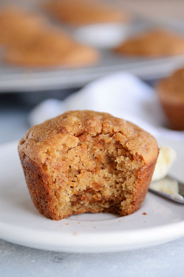 A cooked bran muffin with a bite taken out on a white plate and a pat of butter on a knife next to the muffin.