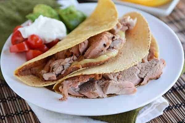 Two corn tortilla pork tacos on a white plate next to sour cream, tomatoes, and avocado.